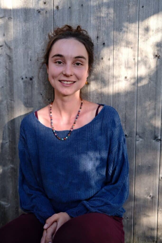 Anuschka Weber lehnt an einer Holzwand und lächelt in die Kamera.